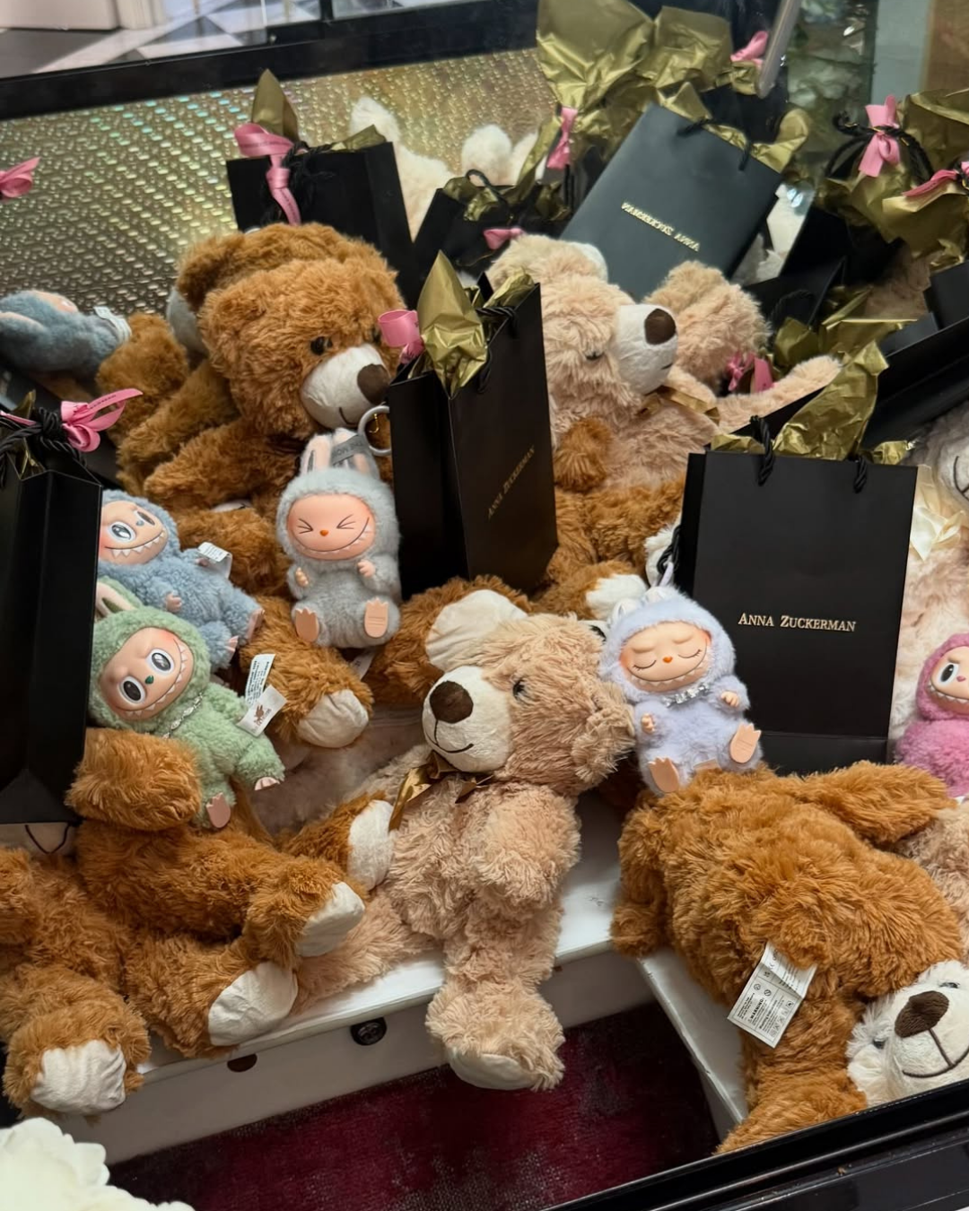 A claw machine filled with plush teddy bears, collectible Labubu figures, and black gift bags from Anna Zuckerman Jewelry with pink ribbon and gold tissue paper.