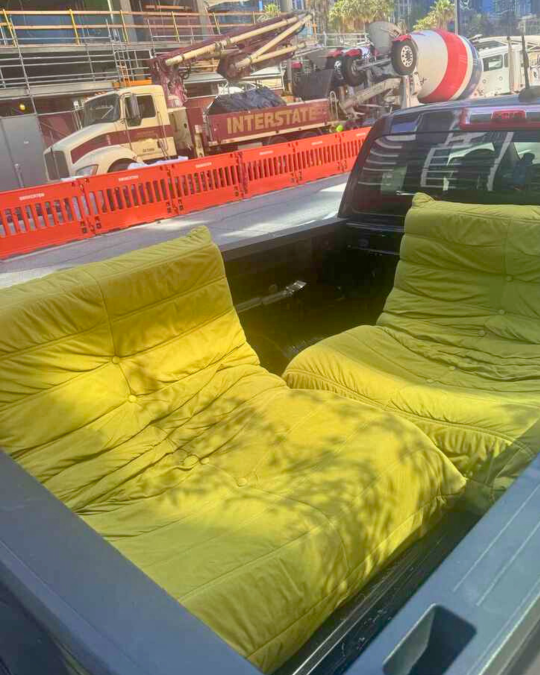 Two chartreuse, Togo-inspired lounge chairs are laid side by side in the bed of a black pickup truck, surrounded by construction barricades and a cement truck in the background.