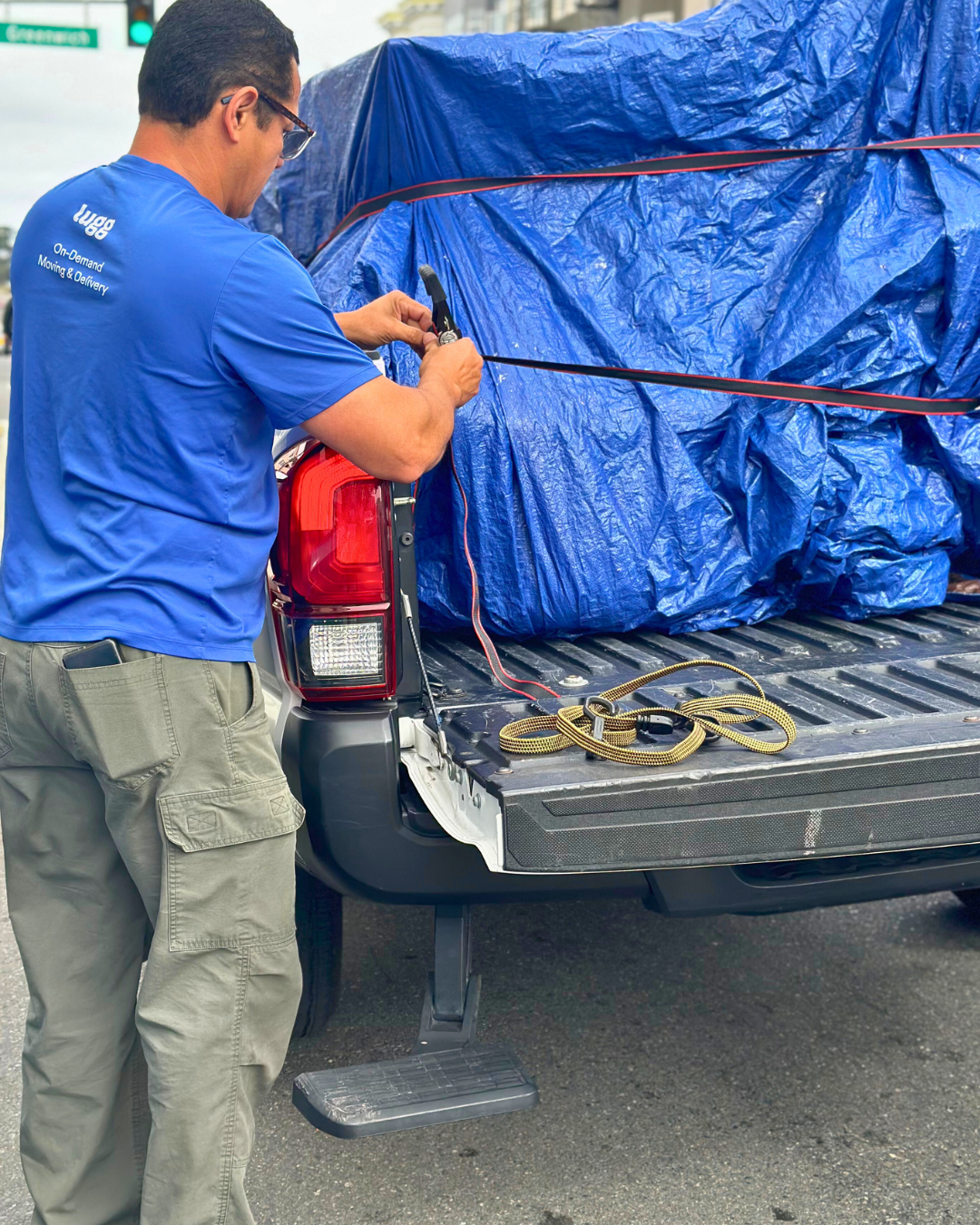 A Lugger in a blue shirt uses a ratchet strap to secure a tarp-covered load in the back of a black pickup truck during a Facebook Marketplace furniture move.