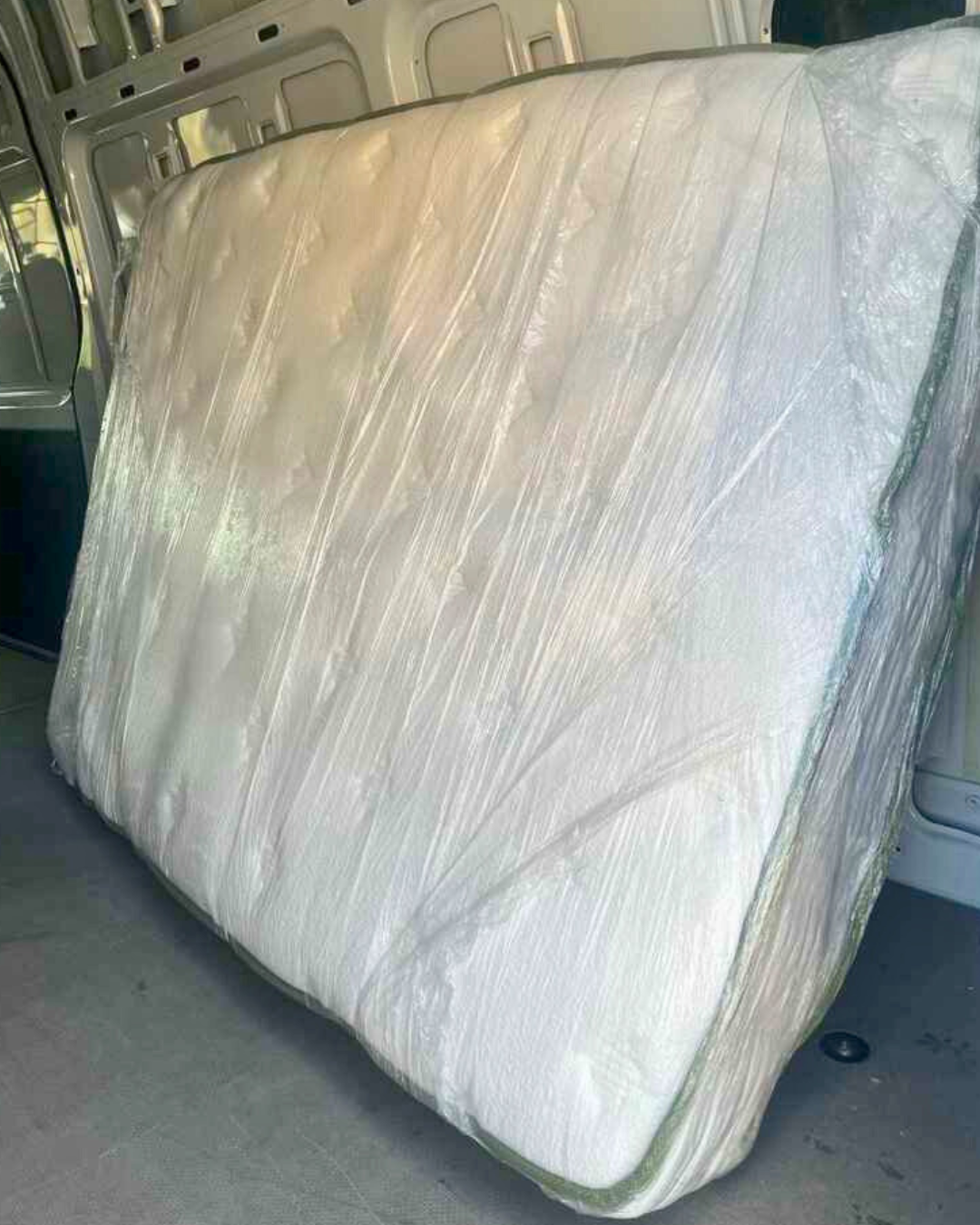 Queen-sized mattress fully wrapped in protective plastic inside the back of a delivery van.