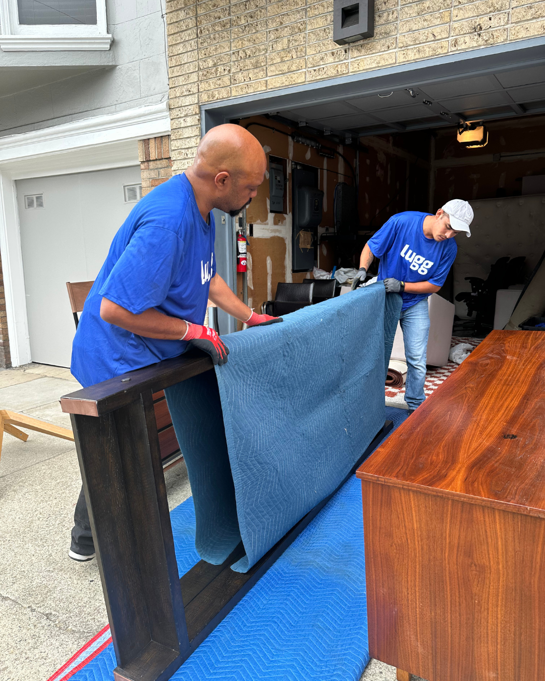 Two Lugg movers placing a moving blanket on a large dresser in a garage.