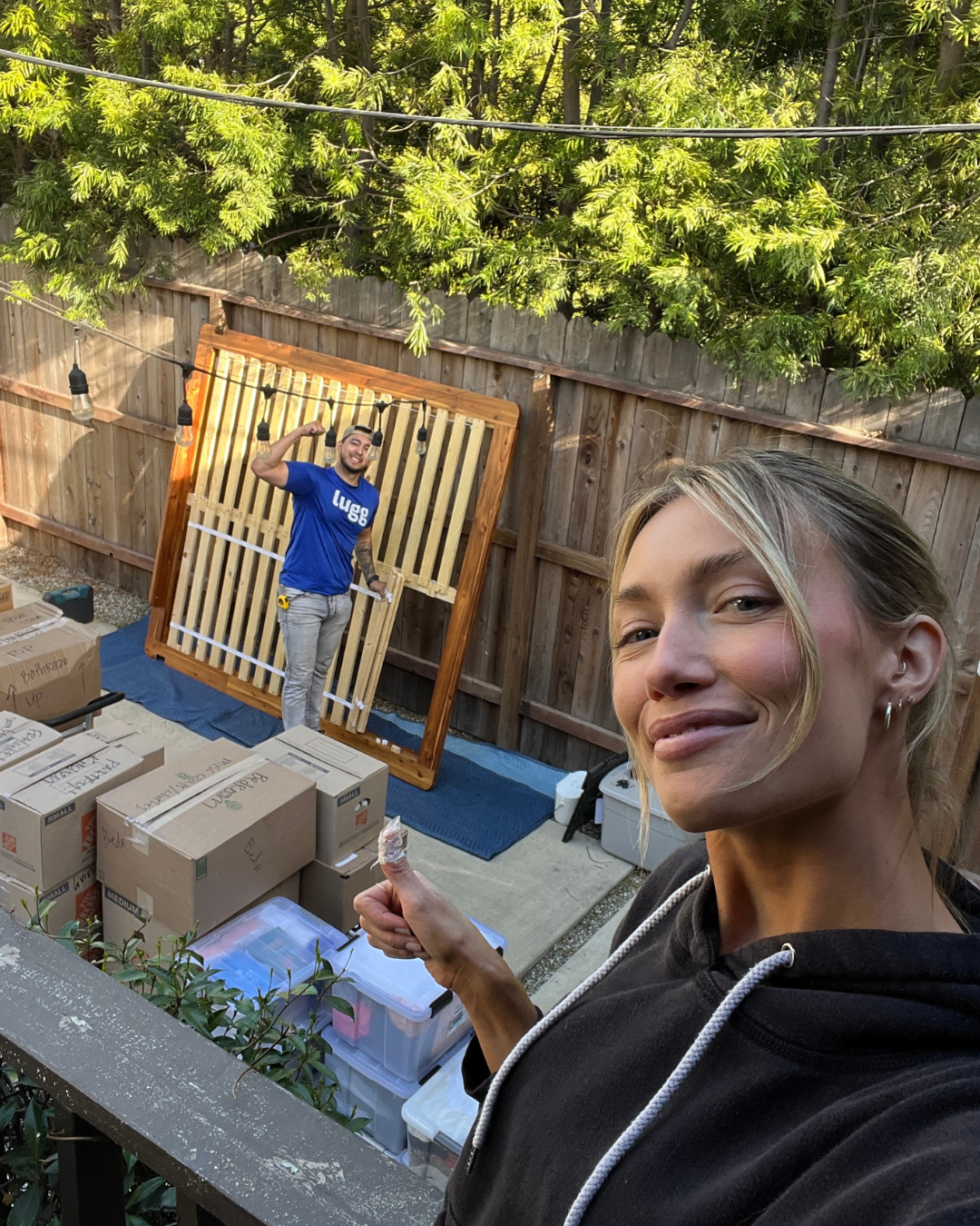 A happy Lugg customer takes a selfie while a mover in a blue Lugg shirt lifts a bed frame in the background.