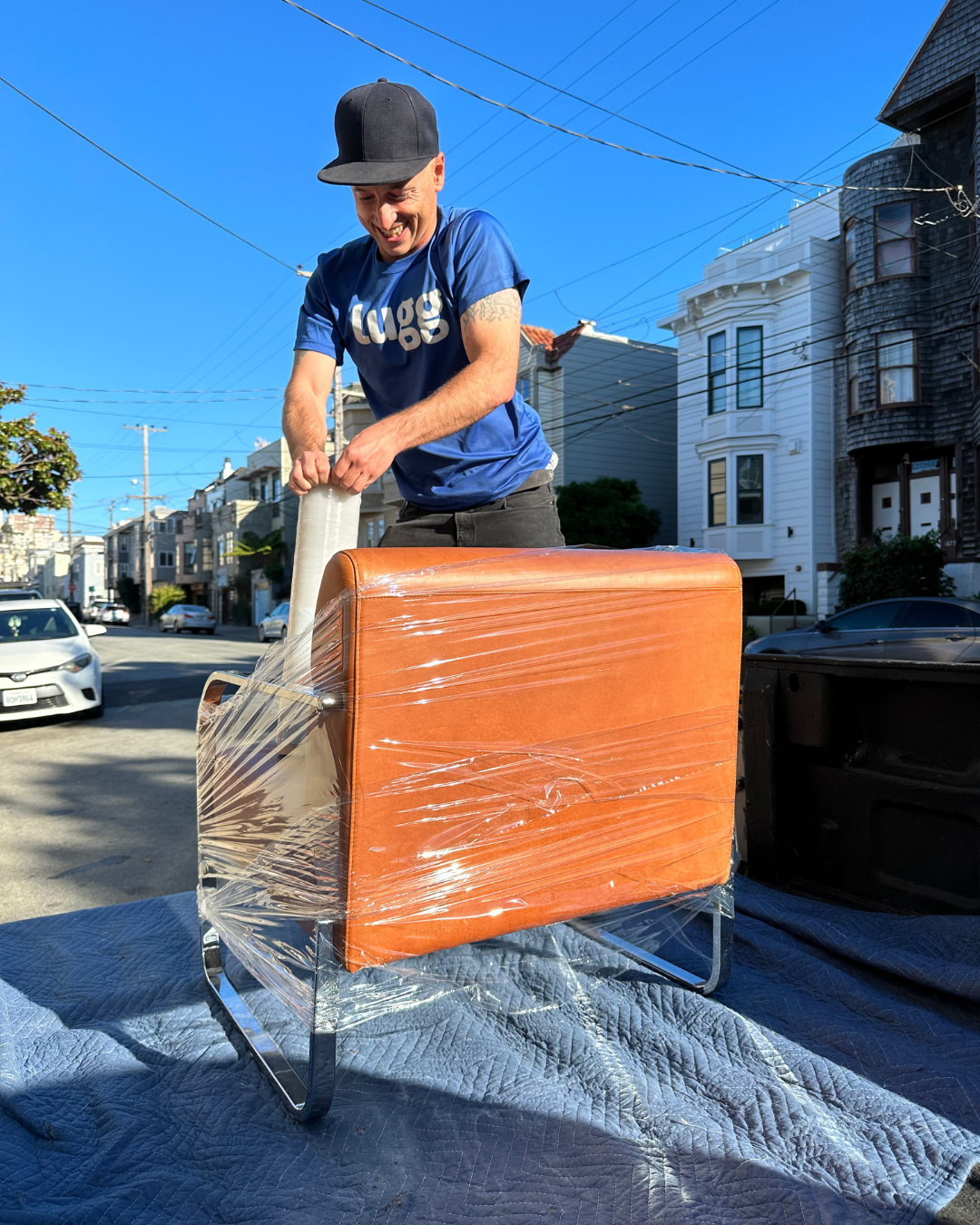 A Lugg mover in a cap wraps an orange chair with plastic wrap on a residential street.