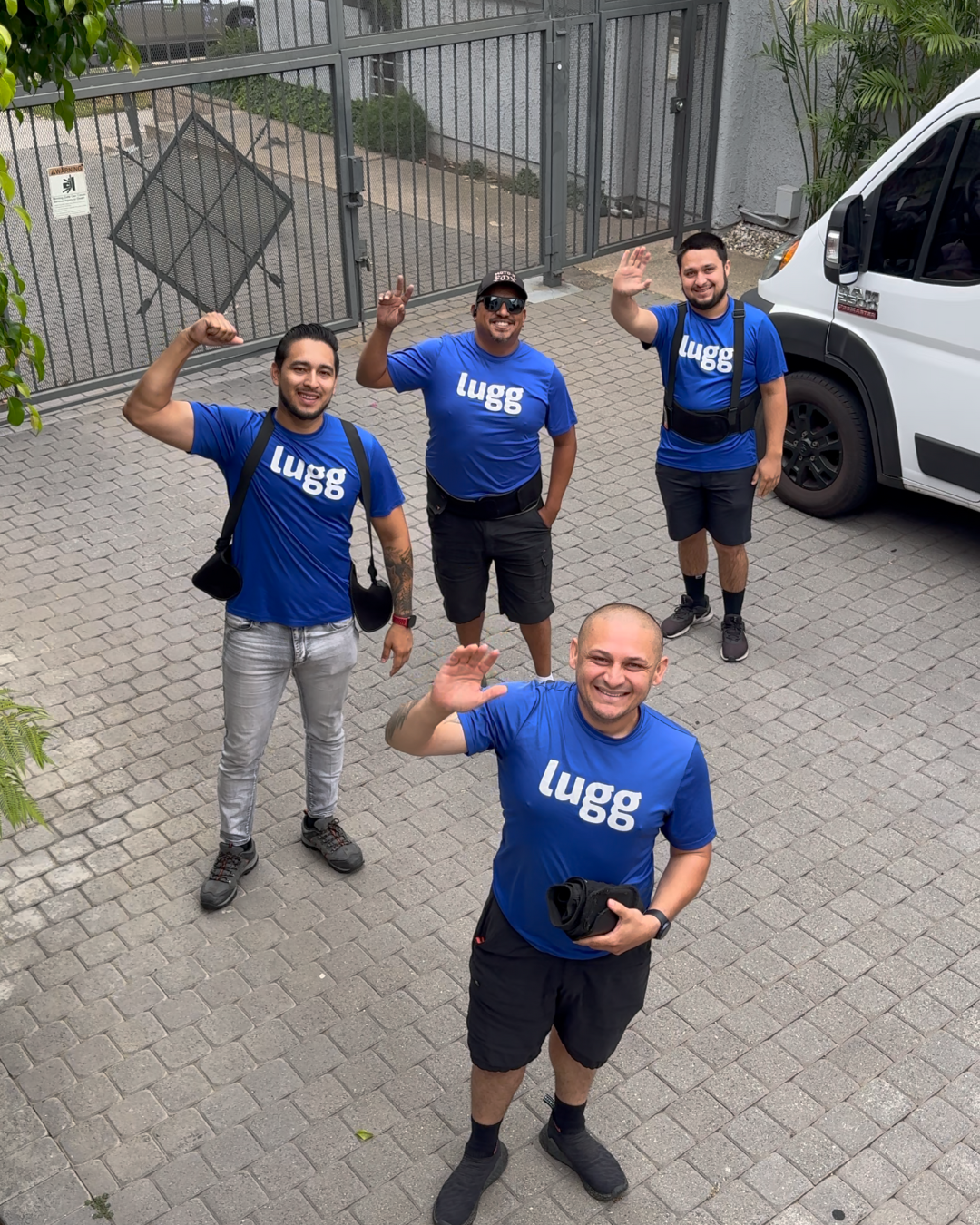 Four Lugg movers in blue shirts waving at the camera in front of a gated entrance.