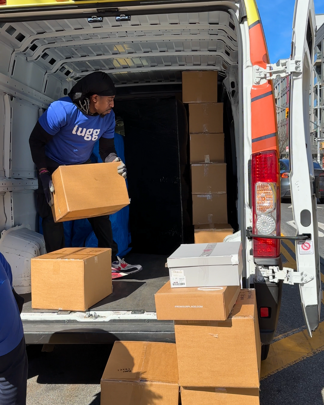 Two movers in blue Lugg shirts rolling a stack of boxes from a van during a same-day move in a Brooklyn neighborhood.