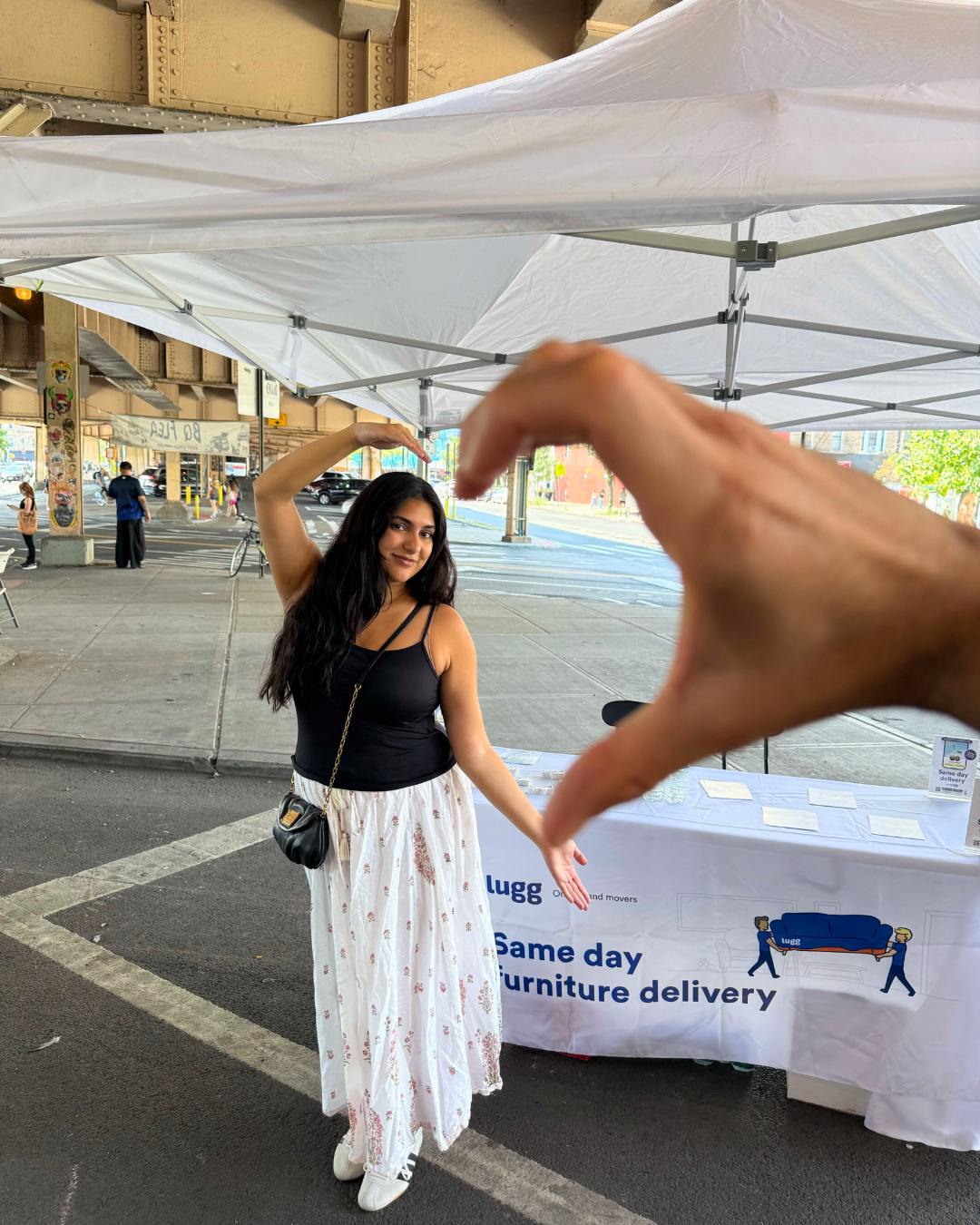 Person posing at the Lugg booth under a white canopy while someone forms a heart shape with their hand in the foreground.
