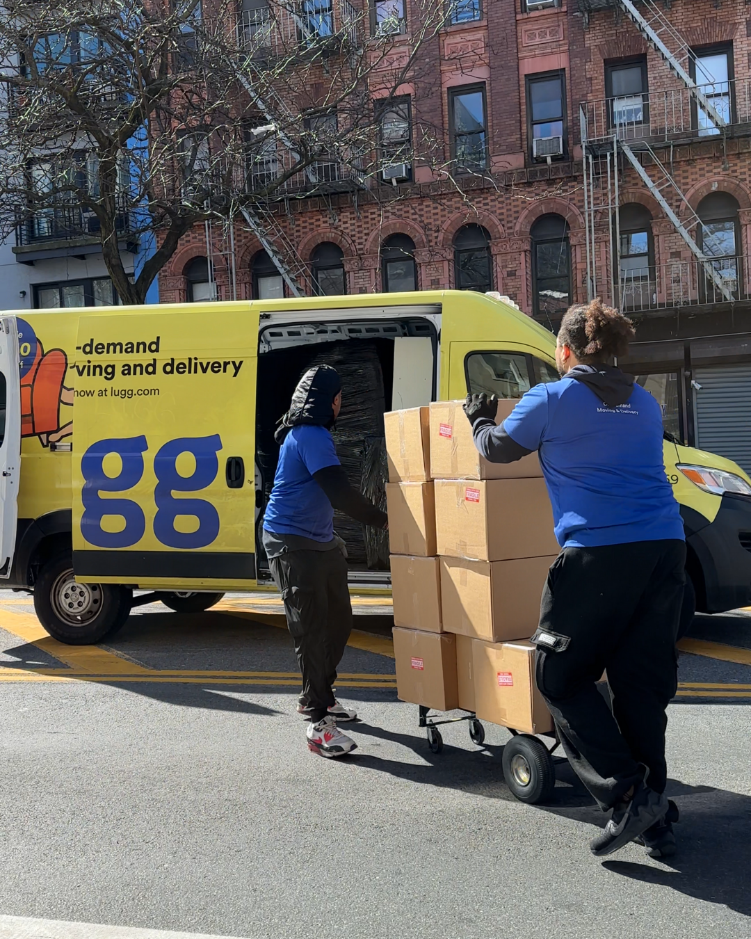 wo movers in blue shirts loading cardboard boxes from a New York City sidewalk into a Lugg van during a same-day move.