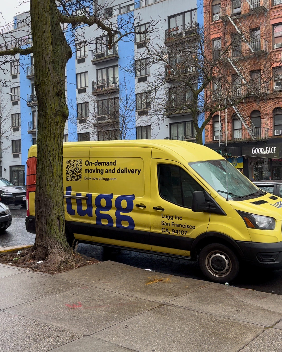 A bright yellow Lugg van parked along a tree-lined street in Brooklyn, surrounded by colorful apartment buildings.