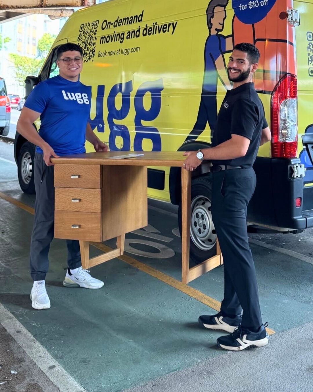 Two Lugg movers carrying a vintage wooden desk in front of a bright yellow Lugg delivery van.