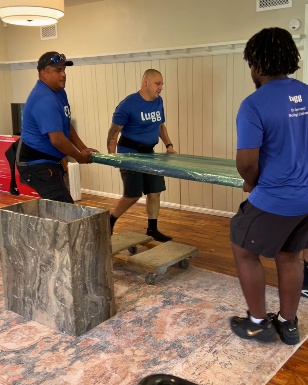 Three Lugg movers in blue shirts lift and wrap large furniture inside a home, demonstrating professional, careful packing support.