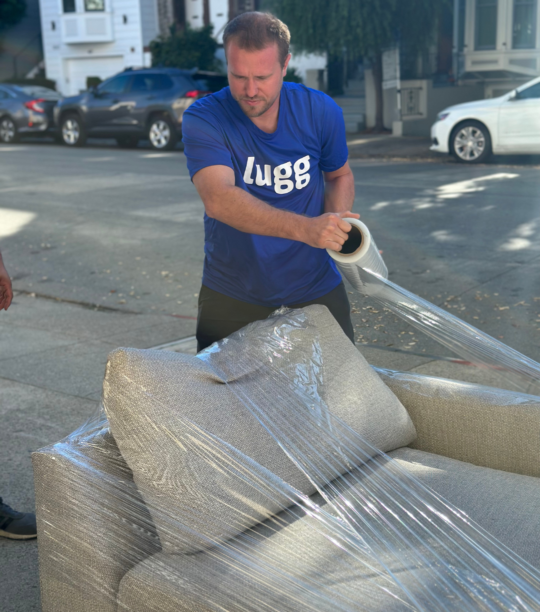 Lugg mover in a blue shirt wrapping a cushioned chair with plastic wrap on a city street.