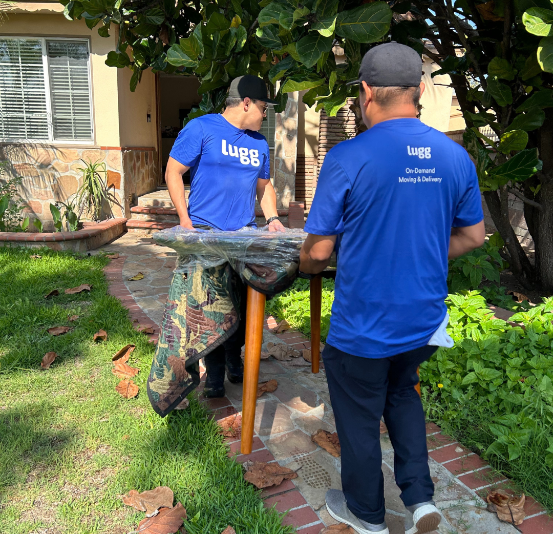 Two Lugg movers in blue shirts carrying a wrapped table along a brick garden path toward a home.
