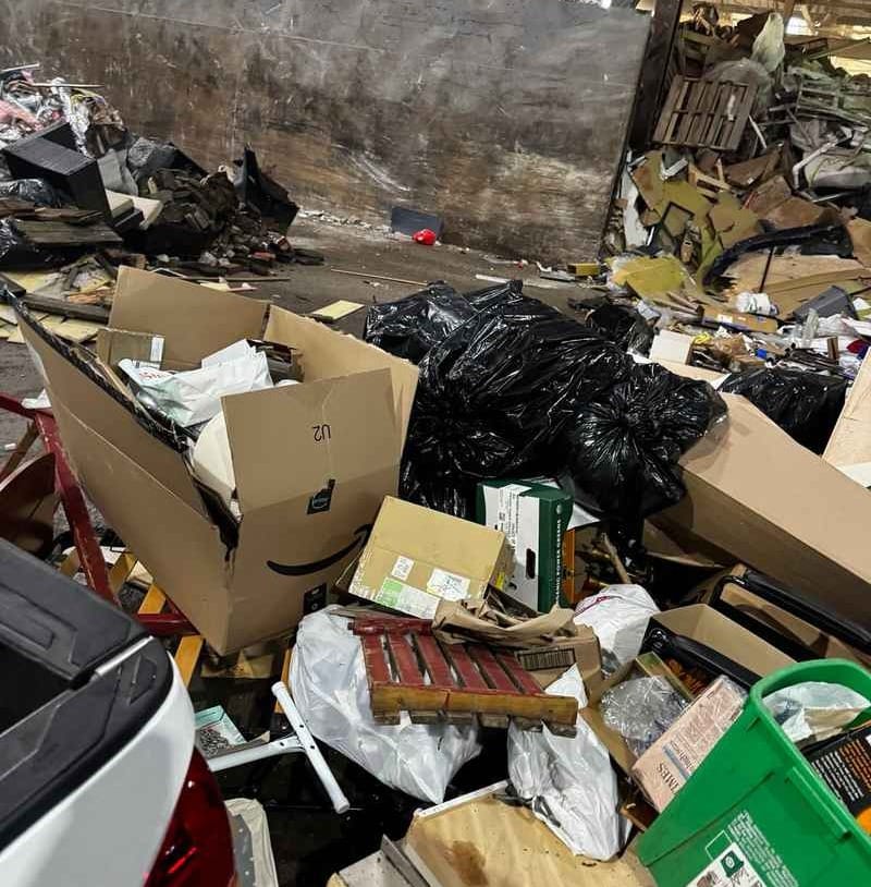 Inside a waste transfer station with piles of household junk, cardboard boxes, and trash bags against an industrial concrete wall