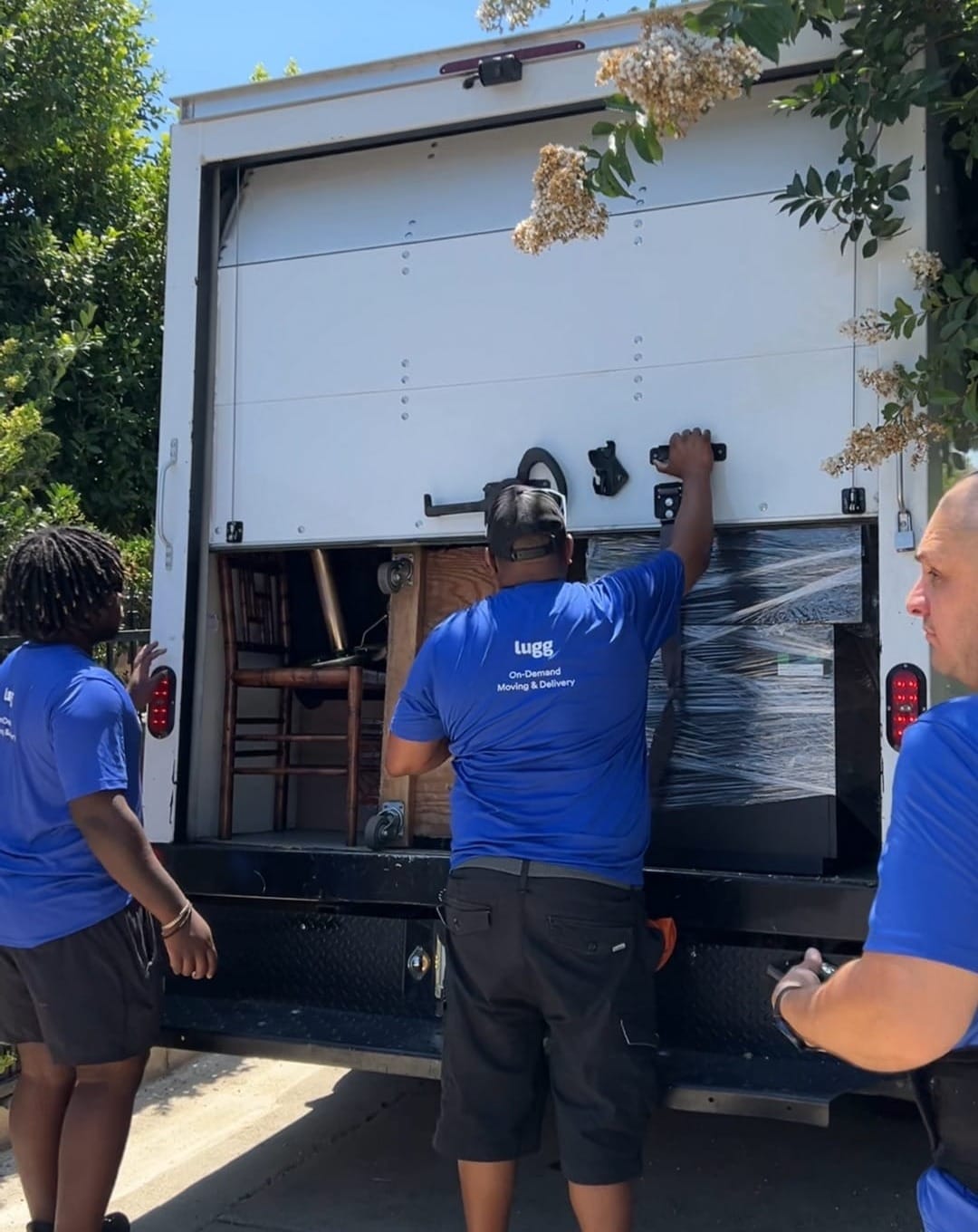 Three Lugg movers in branded blue shirts working together to close up a loaded box truck in a residential neighborhood