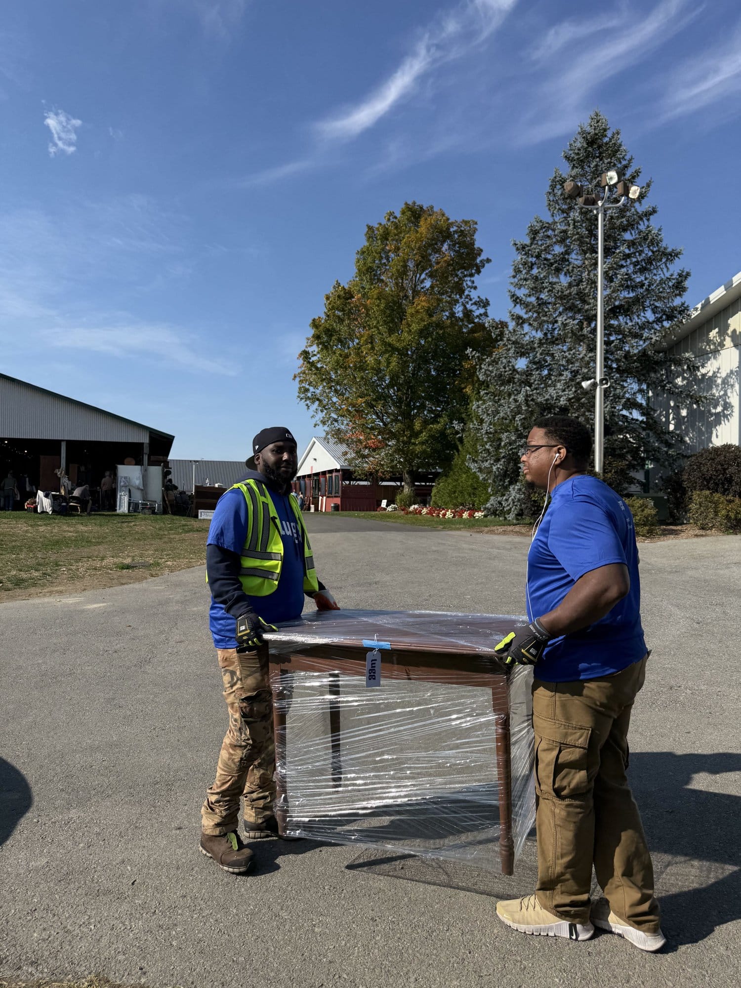 Two Lugg movers carrying a plastic-wrapped antique table across the Dutchess County Fairgrounds