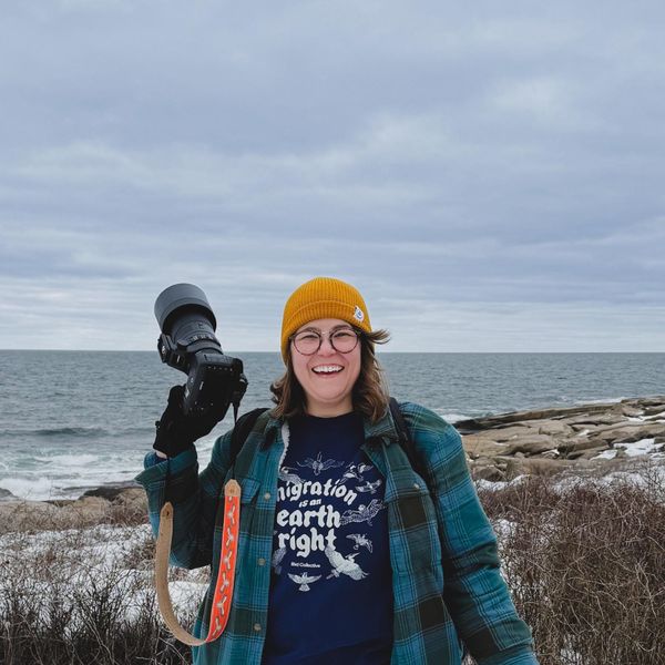 Stephanie smiles on a rocky winter coastline, holding up a camera with a long lens. She wears a yellow beanie, plaid flannel, and a Bird Collective t-shirt.