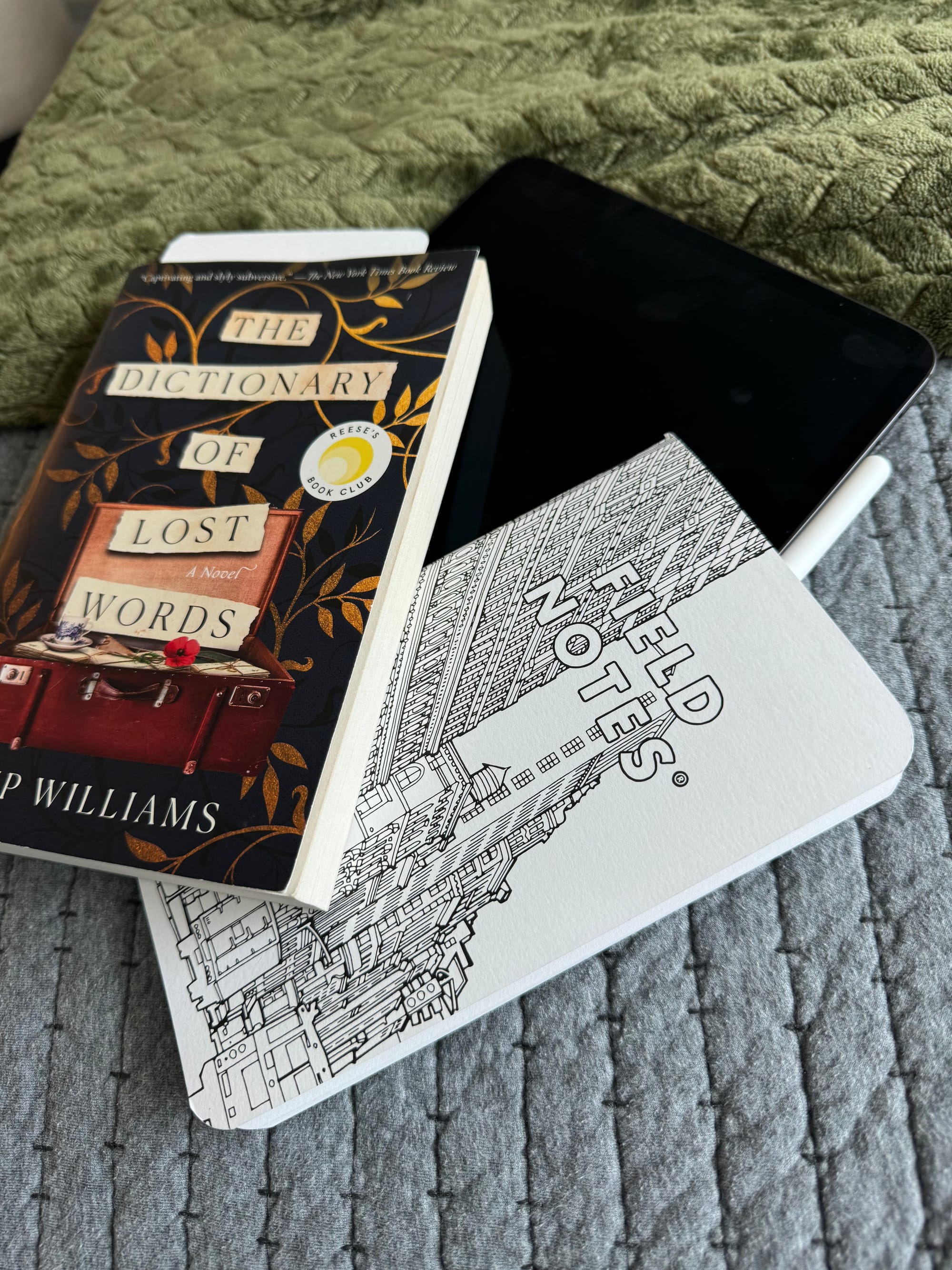 A book, a notebook, and an iPad sit on the edge of a bed