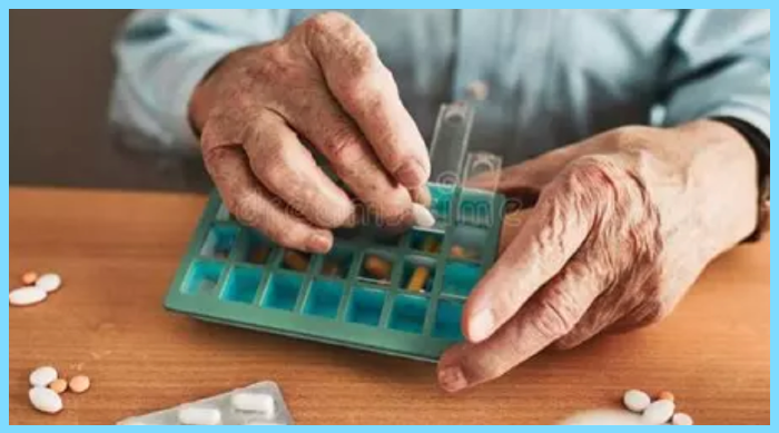 Close up of the hands of an elderly white person filling a daily pill dispenser.  Wearing a light blue shirt, light blue pill dispenser on wooden table surface. Dreamstime.com