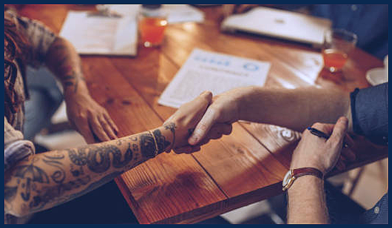 Two people establishing an agreement - showing are the hands and upper arms of two people, one tattooed, shaking hands with paperwork and computers in the background.  istock.com