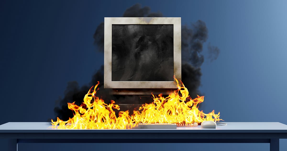 A computer bursting in flames on a desk against a dark blue wall.