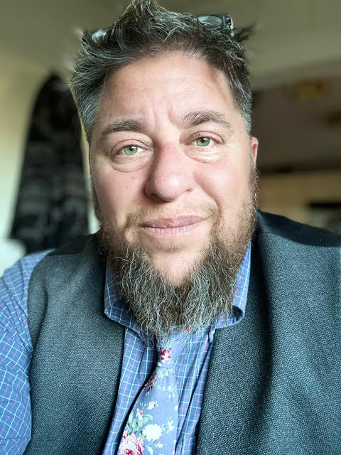 Photo of a white person with a beautiful Ashkenazi face, a greying beard, a vest, a blue stripey shirt and a blue flowerey tie with glasses on his head.