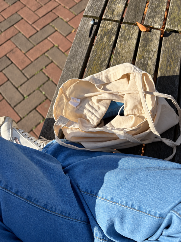 A cream canvas tote on a sunlit wooden bench beside Daphne’s blue jeans and white sneaker.