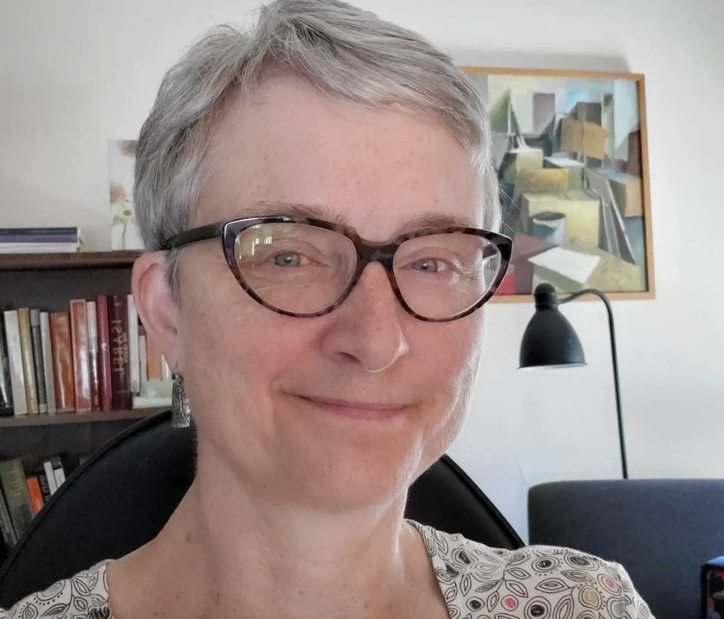 A photo of Liz Lehfeldt, a woman with silver-grey short hair and tortoiseshell glasses.  She is smiling at the camera.