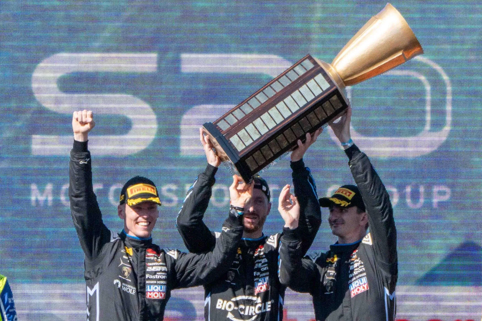 Luca Engstler, Mirko Bortolotti, and Jordan Pepper standing on the Spa 24 Hours podium holding a huge trophy aloft