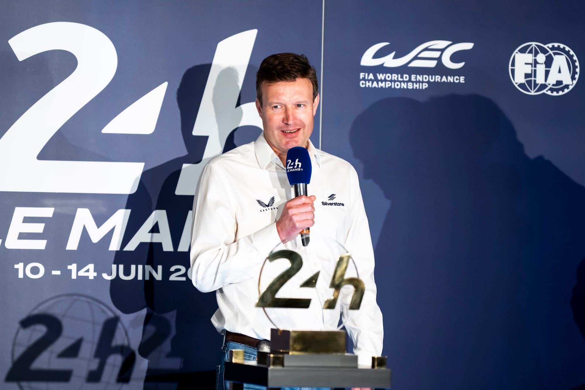 Stuart Pringle, Silverstone's managing director, stands in front of the 24H Le Mans logo, holding a microphone. In front of him is a trophy with the 24H Le Mans logo on it. 
