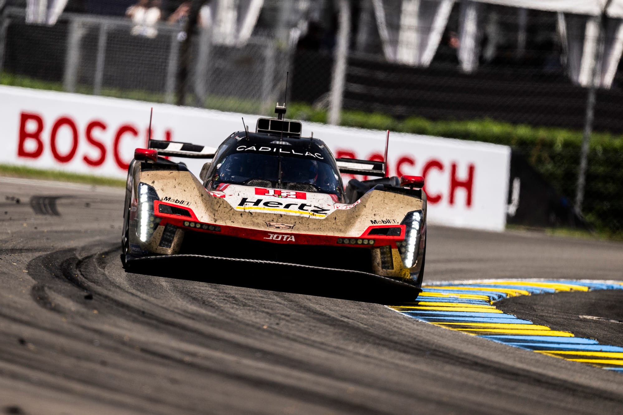 A Cadillac V-Series.R at Le Mans 2026, rounding a corner with the left-hand side tyres partially on a blue and yellow kerb