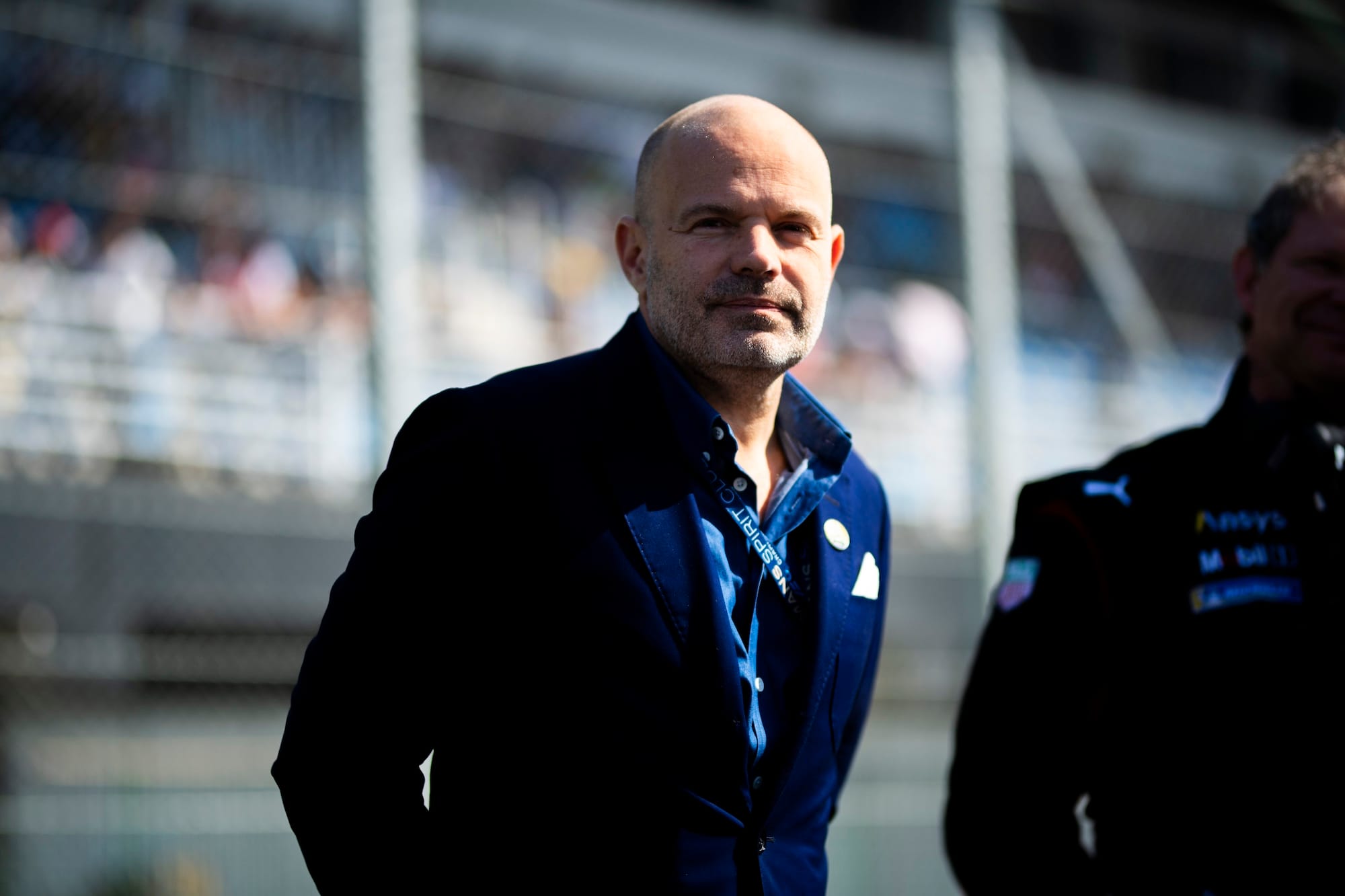 Frederic Lequien at a race track in a dark blue suit, hands behind his back, smiling and looking at the camera