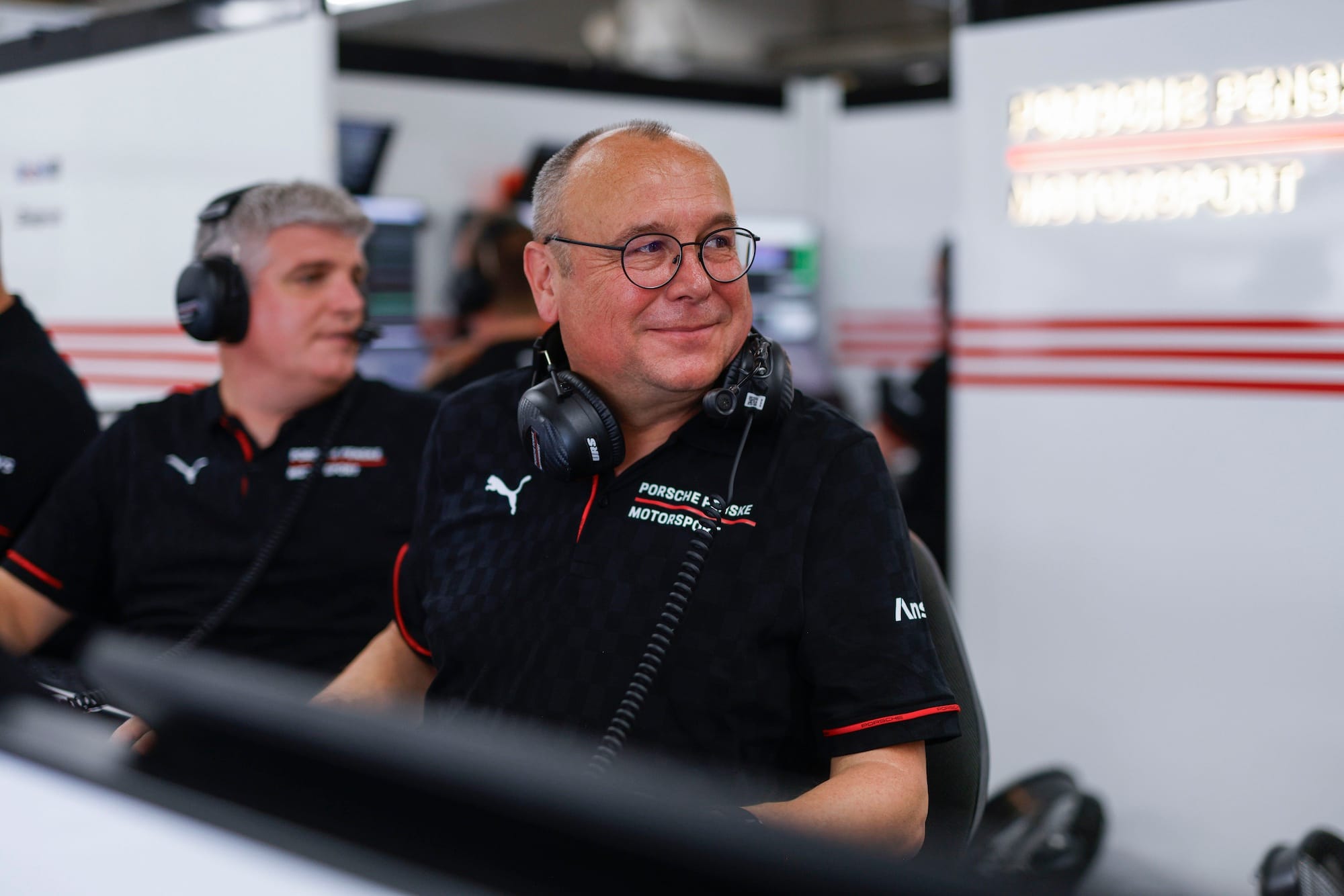 Porsche's Urs Kuratle in the Porsche garage, with headset round his neck, smiling and glancing to his left.