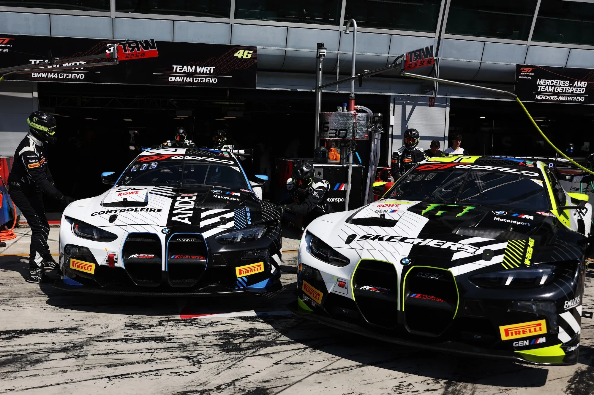 Two BMW M4 GT3 Evos in the pits, some crew members around them, with the cars raised on wheeled dollies