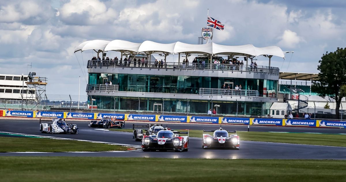 The two Toyota TS050s round Luffield corner at Silverstone, joined by other prototypes behind. The BRDC clubhouse is in the background.