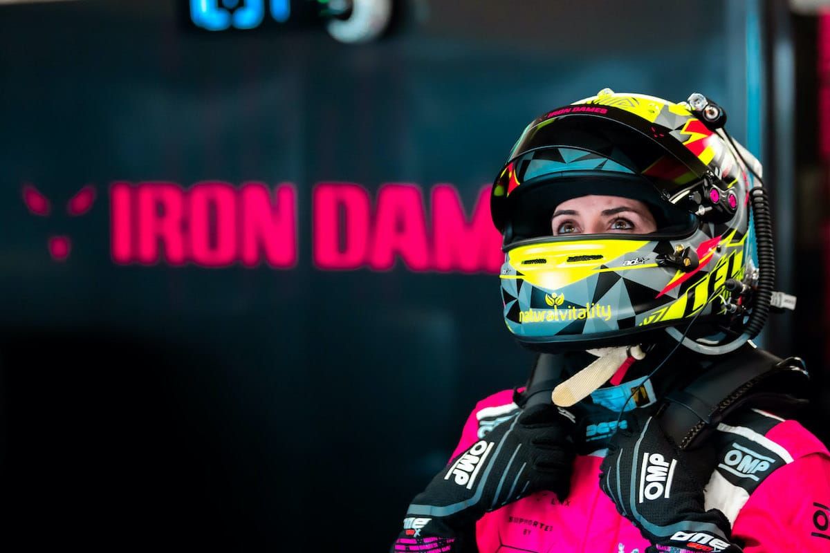 Celia Martin looks up in her racing suit and helmet with the Iron Dames logo in the background