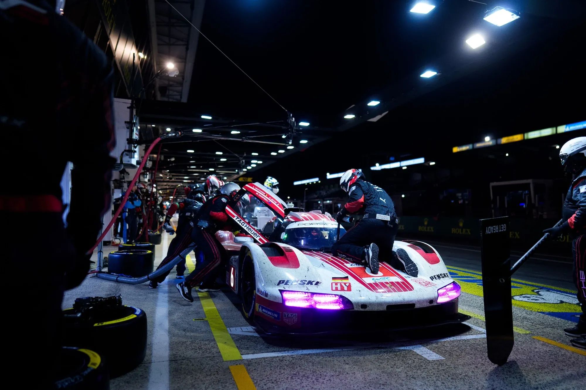 The #6 Porsche in the pits at night at Le Mans, with the car being refueled and a mechanic cleaning the windscreen