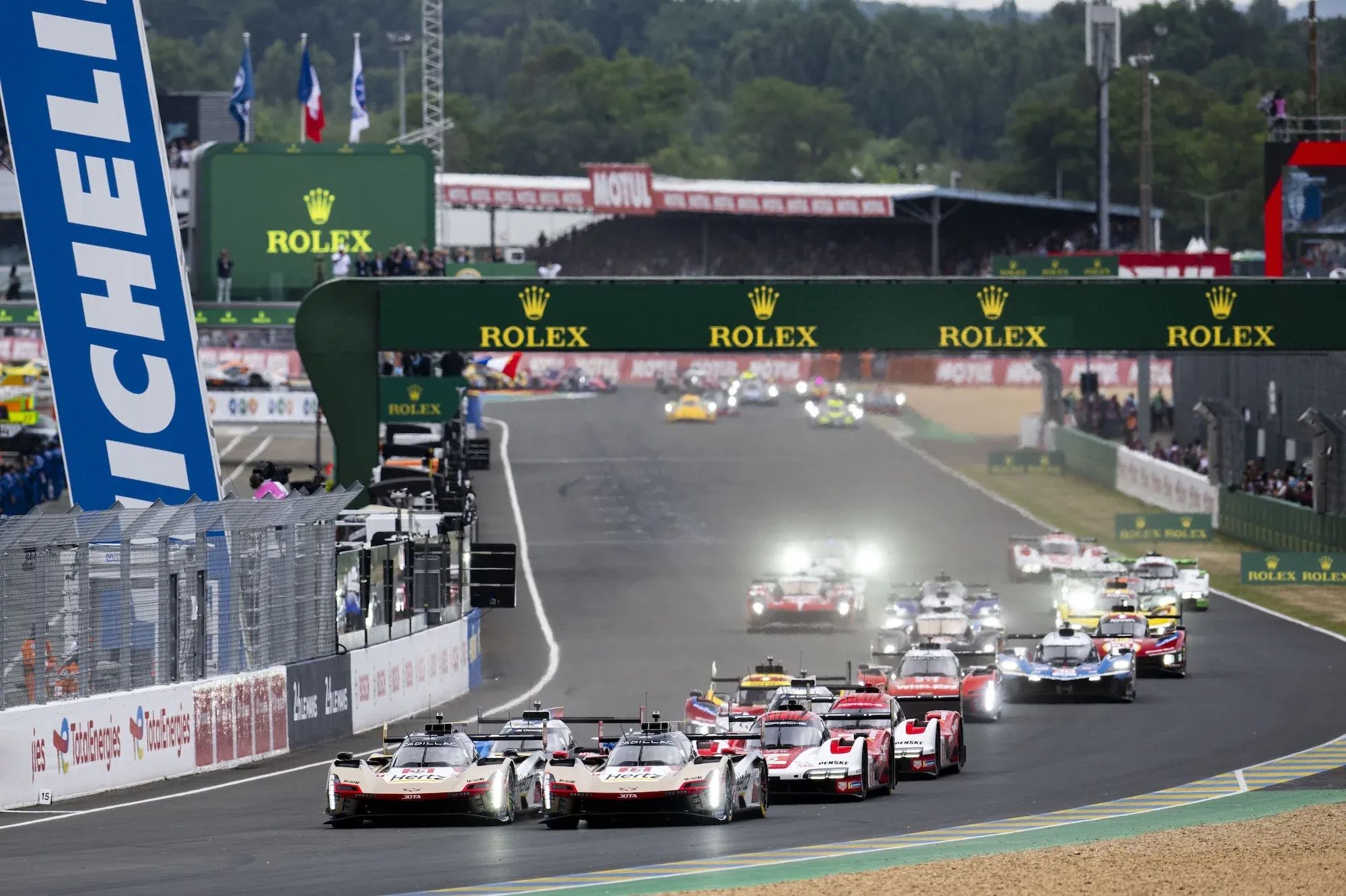 Start of the race, Le Mans 2025, with the cars passing the main straight with the pit lane in the background on the left