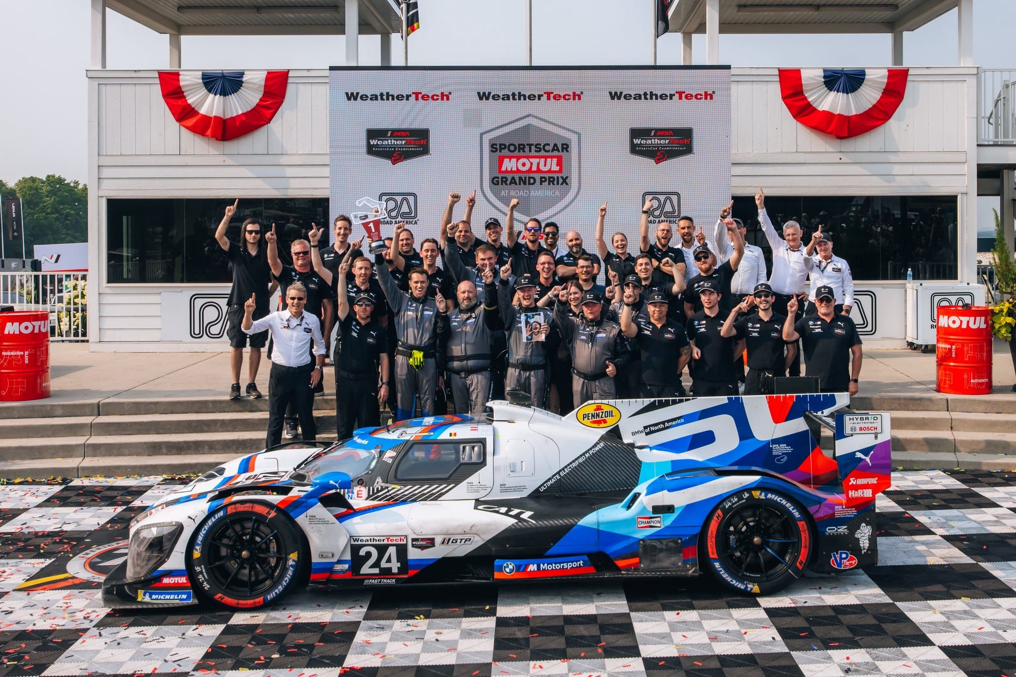 The winning #24 BMW M Hybrid V8 at Road America, with the team crew cheering behind the car