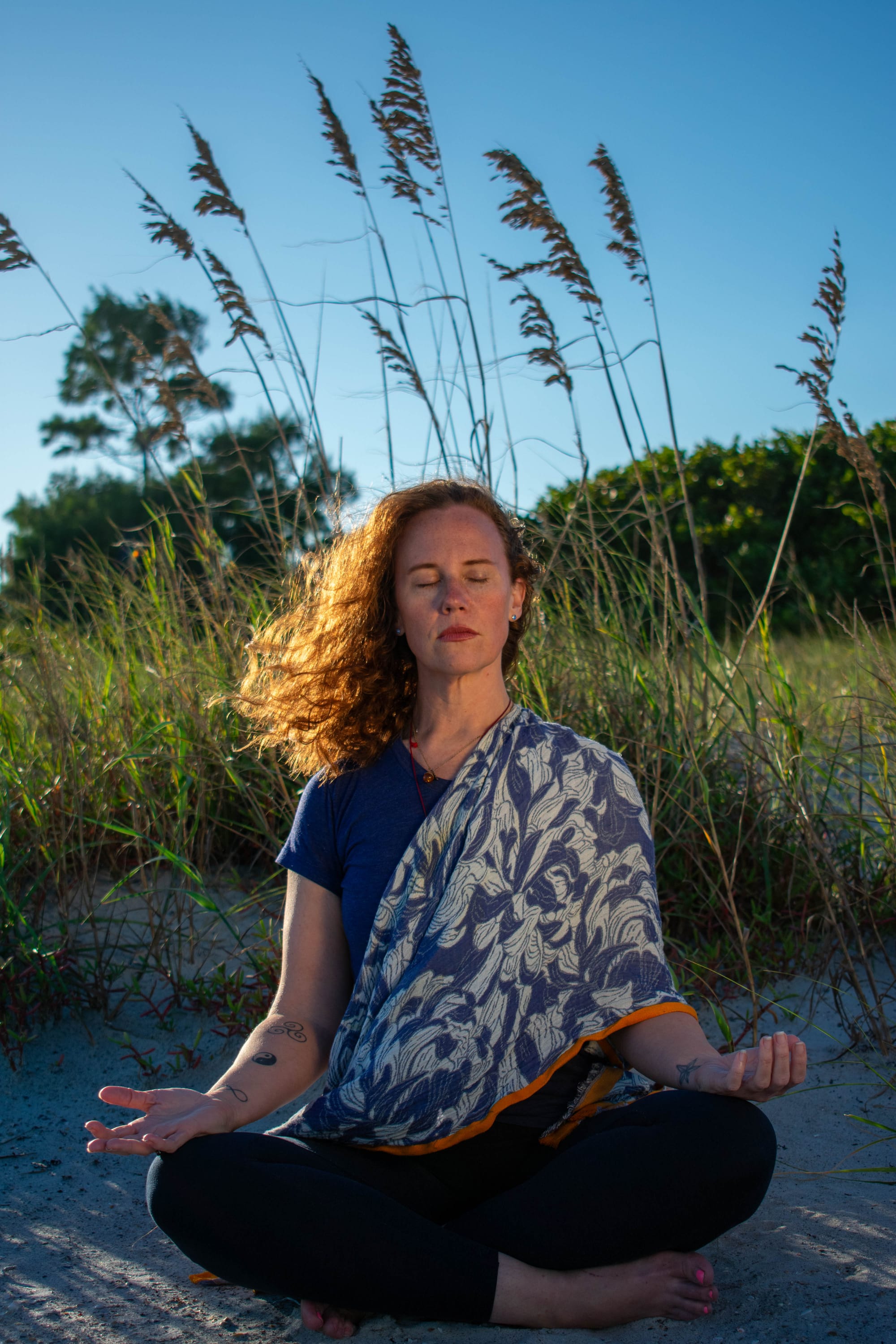 A red-haired woman sitting crosse legged in the sand with eyes closed in meditation with sea grasses and a warm blue sky background