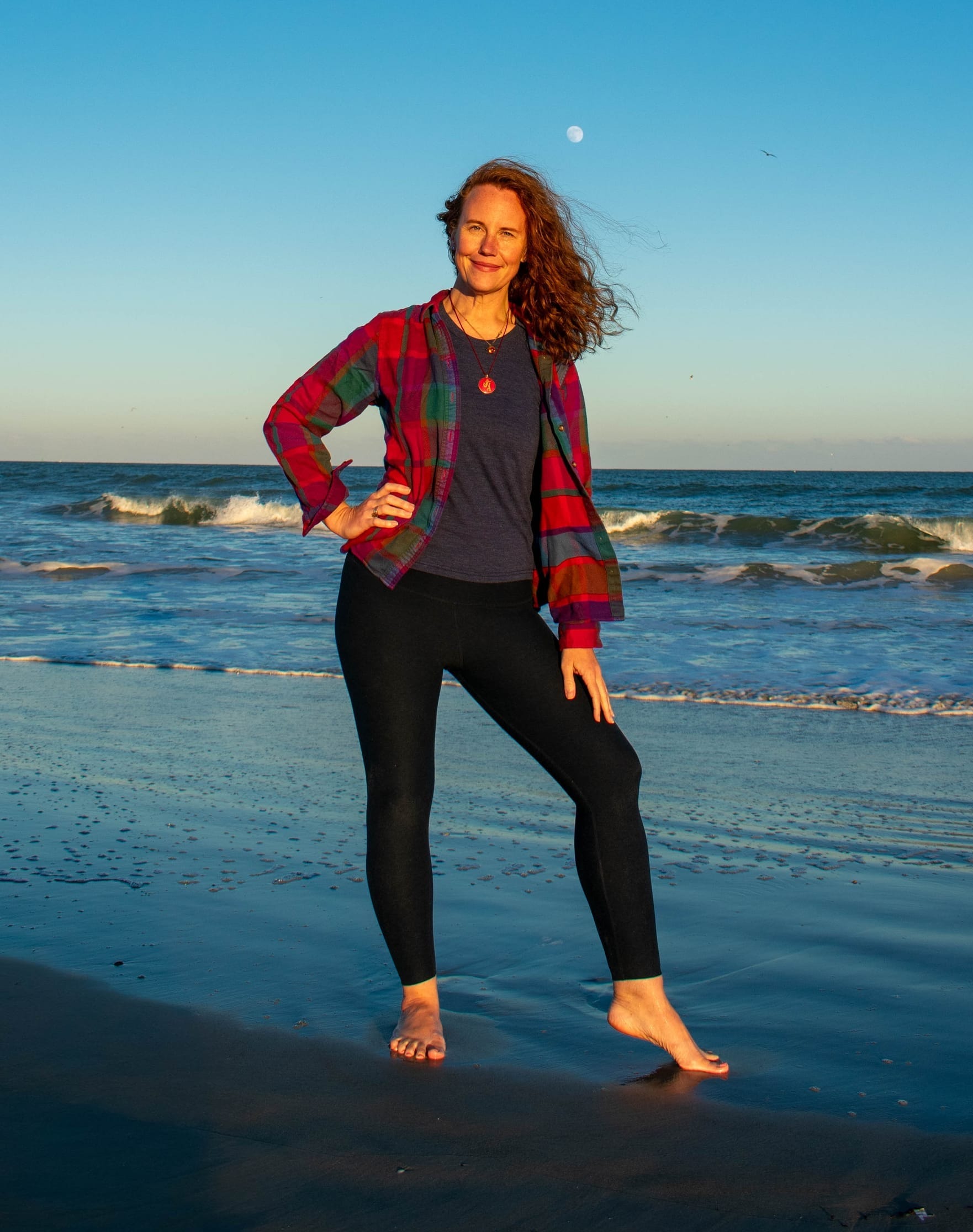 A woman with curly red hair in dark leggings and a bright flannel shirt standing in wet sand on the shore with a bright blue ocean and sky in the background