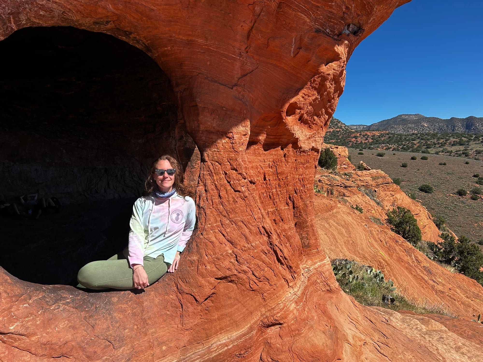 A woman with sunglasses and a tie-dyed shirt and green leggings sits in a hole of a red rock formation with desert scrub landscape in the background