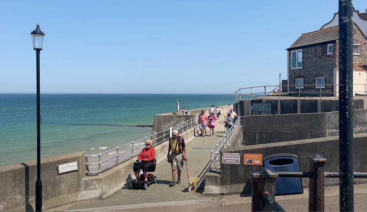 older man walking dog next to older woman on a mobility scooter along the concrete promenade at Sheringham.