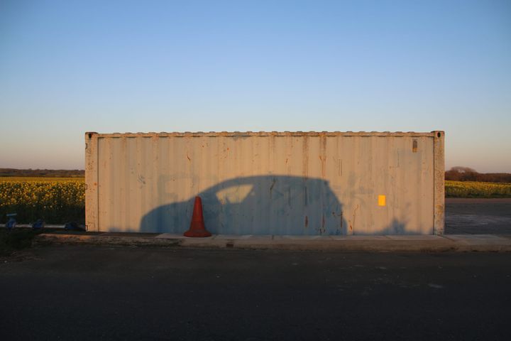 The shadow of a car against a shipping container, with fields and sky in the background