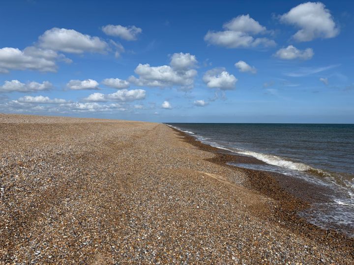 Photos with shingle beach on the left and the sea on the right, with white fluffy clouds in a blue sky