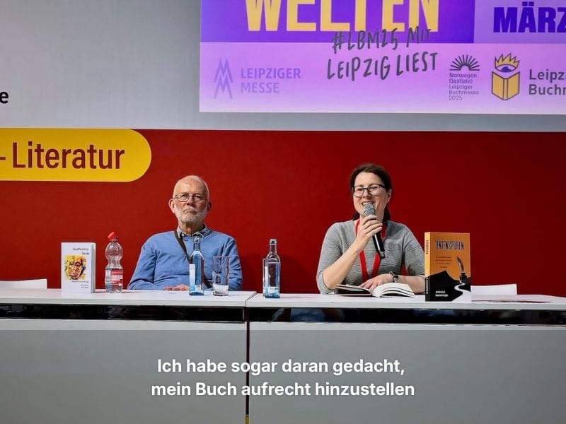 Andrea mit Mikrofon auf der Bühne, liest aus ihrem Buch „Tintenspuren“, neben ihr sitzt der Autor Joachim Oelßner.