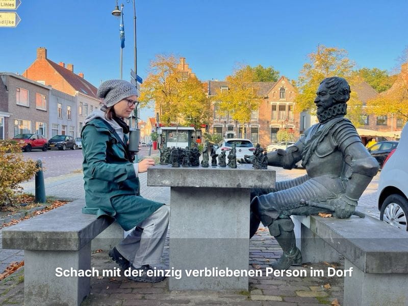 Andrea sitzt auf einem Marktplatz in einem niederländischen Dorf und spielt bei Sonnenschein im Herbst Schach mit einer Bronzestatue.
