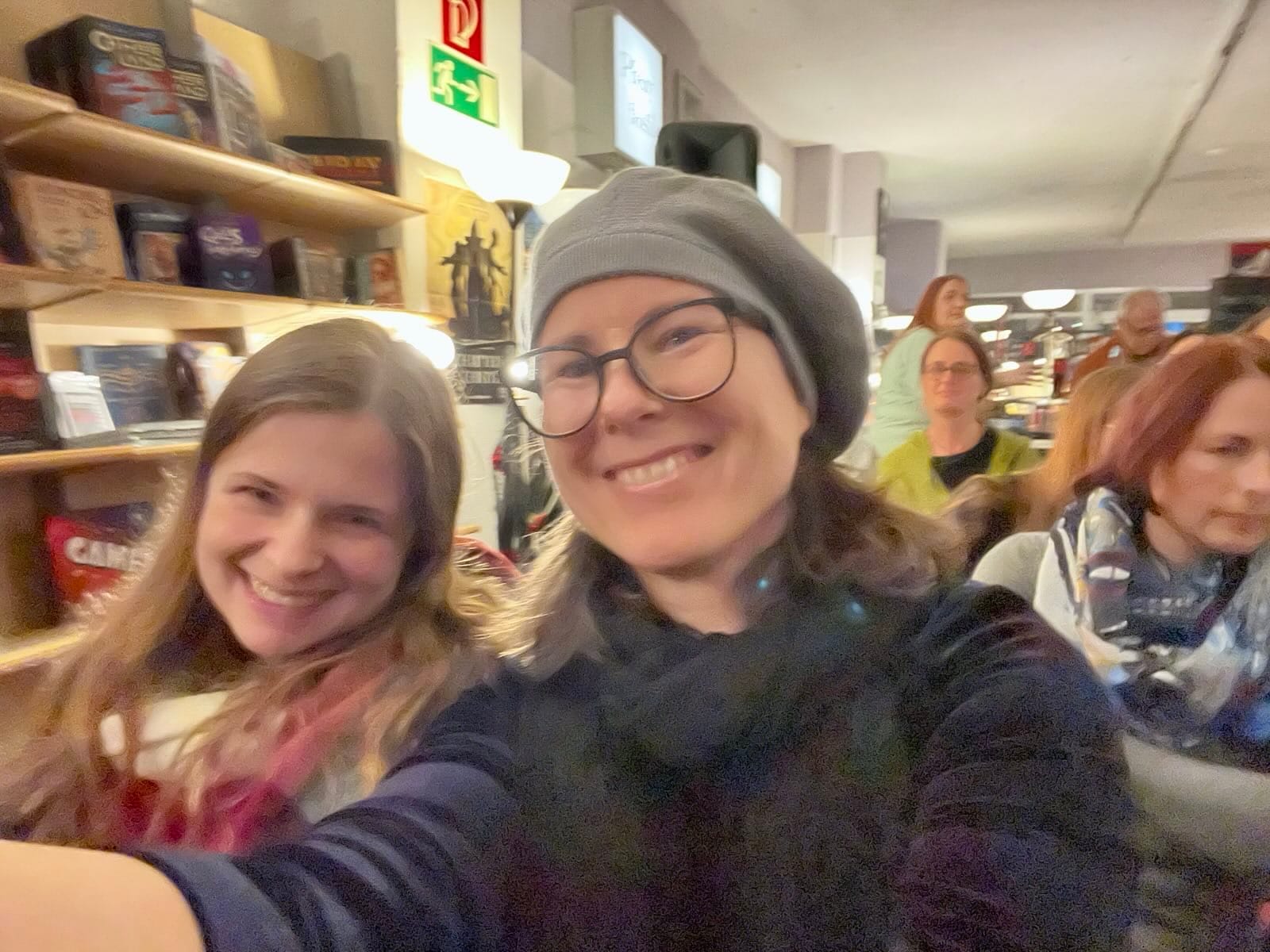 Andrea und ihre Autorinnenkollegin Sascha lachend auf einer Lesung im Drachenwinkel, mit Buchladen und Publikum im Hintergrund