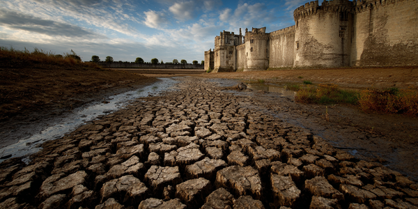 A medieval castle with a completely dried and cracked moat, leaving exposed mud and fissures where water once surrounded the fortress.