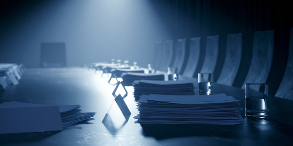 An empty conference table stretches into darkness, one side lined with stacks of briefing documents, name placards, and glasses of water. No one has arrived.