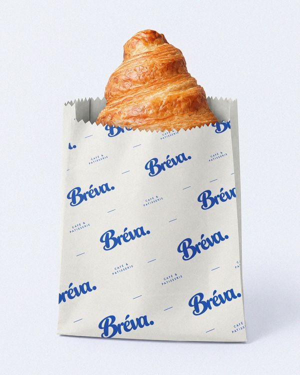 Cream croissant paper sleeve mockup with repeating blue Bréva Café & Pâtisserie branding and a freshly baked croissant on a clean white background.